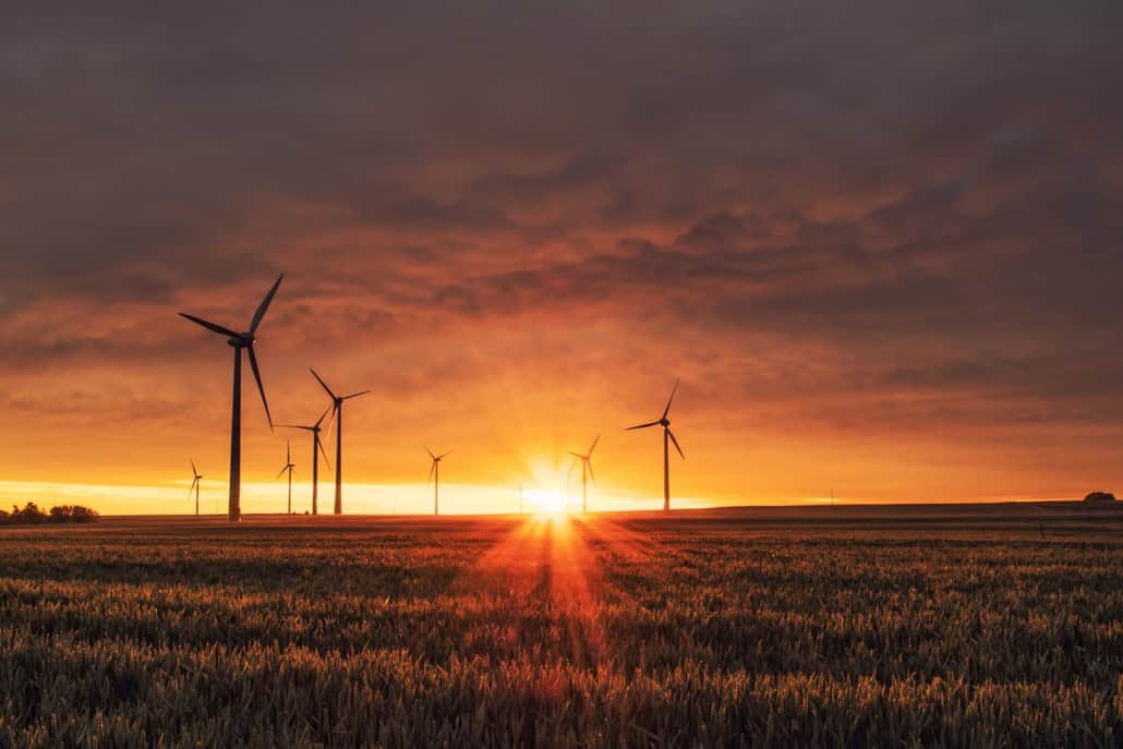 Wind turbines in a field at sunset, with the sun shining through clouds and casting an orange glow.