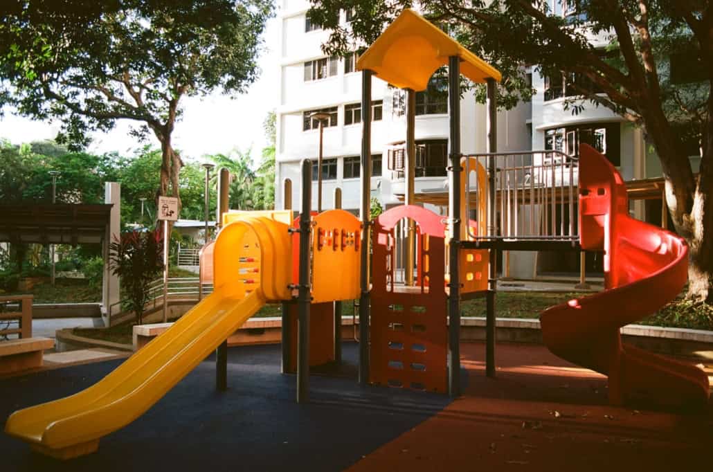 Colourful playground with yellow slide and red spiral slide, set outdoors near block of flats and trees.