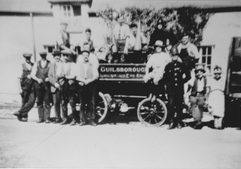 Black-and-white photo of a group of people standing around an old vehicle labelled "Guilsborough.