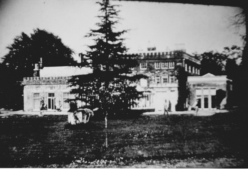 Black-and-white photo of a large, old mansion with trees and lawn in the foreground.